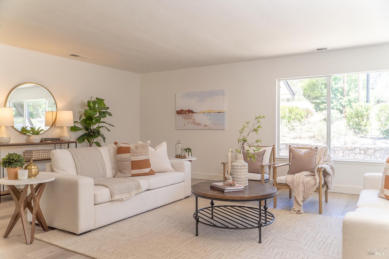 This bright and inviting living room features a neutral color palette with a comfortable cream-colored sofa, two accent chairs, and a round wooden coffee table. A large window allows for ample natural light, while a circular mirror and abstract wall art add decorative interest to the space. The room is finished with a textured area rug and a potted plant, creating a warm and cohesive atmosphere.