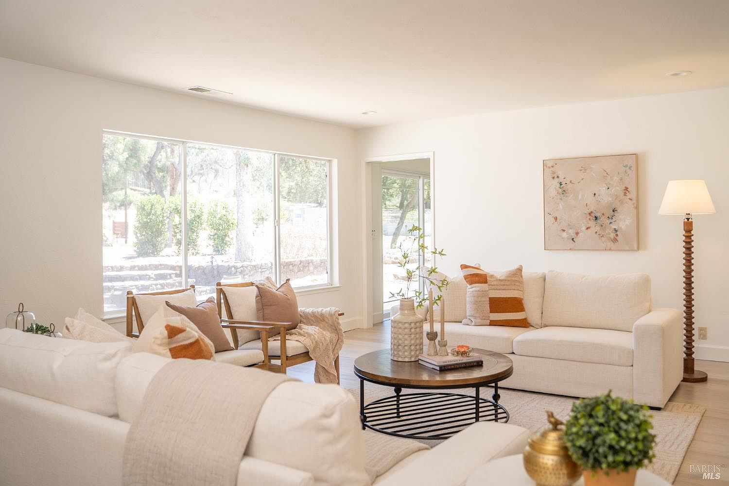 This bright and airy living room features a neutral color palette with comfortable white sofas and two accent chairs arranged around a round wooden coffee table. Large windows allow ample natural light to fill the space, providing a view of the outdoor greenery, while a tall floor lamp and abstract wall art add warmth and character. The room is designed with a clean, modern aesthetic that feels both inviting and spacious.