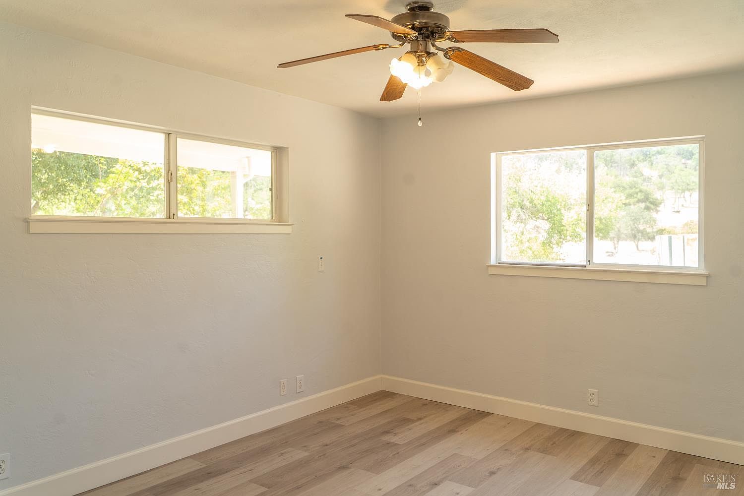 This bright and neutral bedroom features light-toned wood-look flooring and soft, pale-painted walls. Two large windows allow for ample natural light, while a ceiling fan with integrated lighting provides overhead illumination. The room offers a clean, minimalist aesthetic, serving as a versatile blank canvas for any decor style.