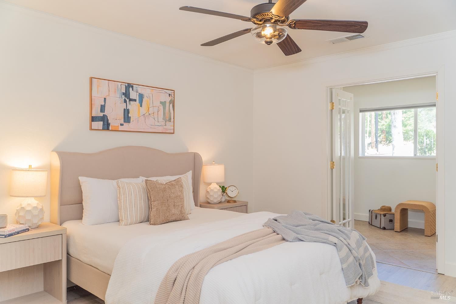 This bright and airy bedroom features a neutral color palette with a plush upholstered headboard and layered bedding in soft whites and tans. A modern ceiling fan hangs above the bed, while a piece of abstract art adds a touch of color to the wall. Through the open French doors, a secondary space is visible, creating an inviting and open-concept feel.