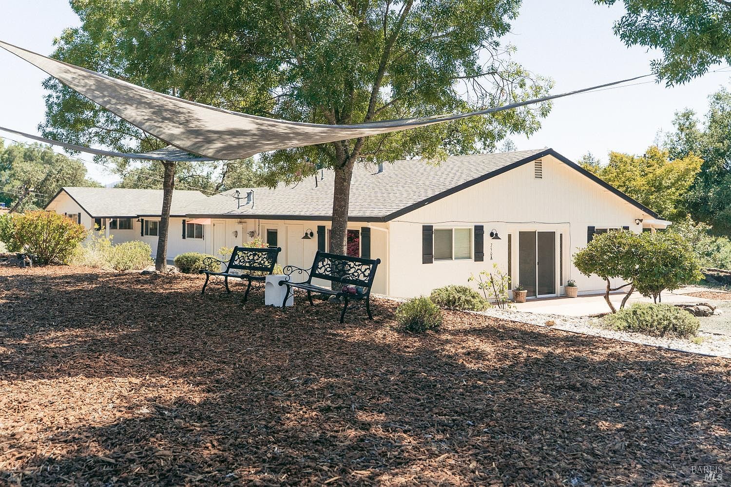 This exterior view showcases the rear of a single-story home with light-colored siding and a dark shingled roof. A large shade sail is suspended over a mulched area featuring two black metal benches, creating a comfortable outdoor seating space beneath a mature tree. The home includes a sliding glass door leading to a patio, with surrounding landscaping that provides a peaceful, private atmosphere.