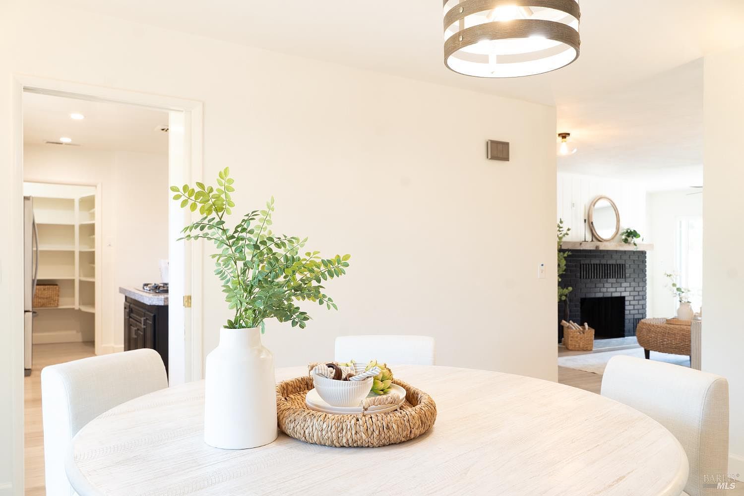 This bright dining area features a round, light-toned wood table centered under a modern drum-style pendant light. A woven tray holds a decorative bowl and greenery, while the open floor plan provides a clear view into a kitchen with pantry storage and a living area with a dark-painted brick fireplace. The neutral color palette and clean lines create an inviting, airy atmosphere suitable for casual or formal dining.