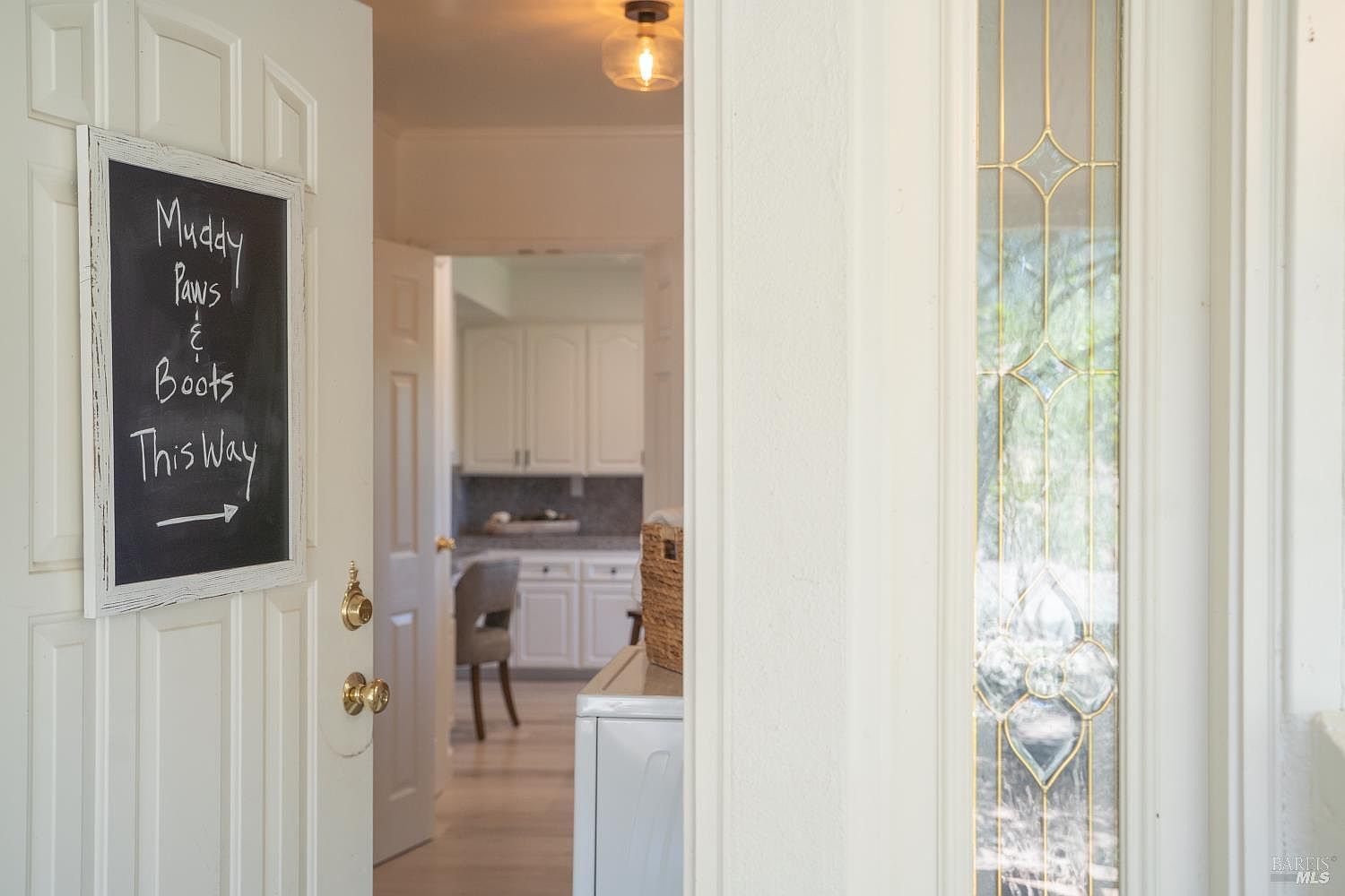 This entryway view captures a white door featuring a charming chalkboard sign that reads 'Muddy Paws & Boots This Way' with an arrow pointing toward the interior. Through the open doorway, a glimpse of a bright, clean kitchen with white cabinetry and a grey chair is visible. To the right, a decorative glass panel with a floral motif adds a touch of classic elegance to the transition between spaces.