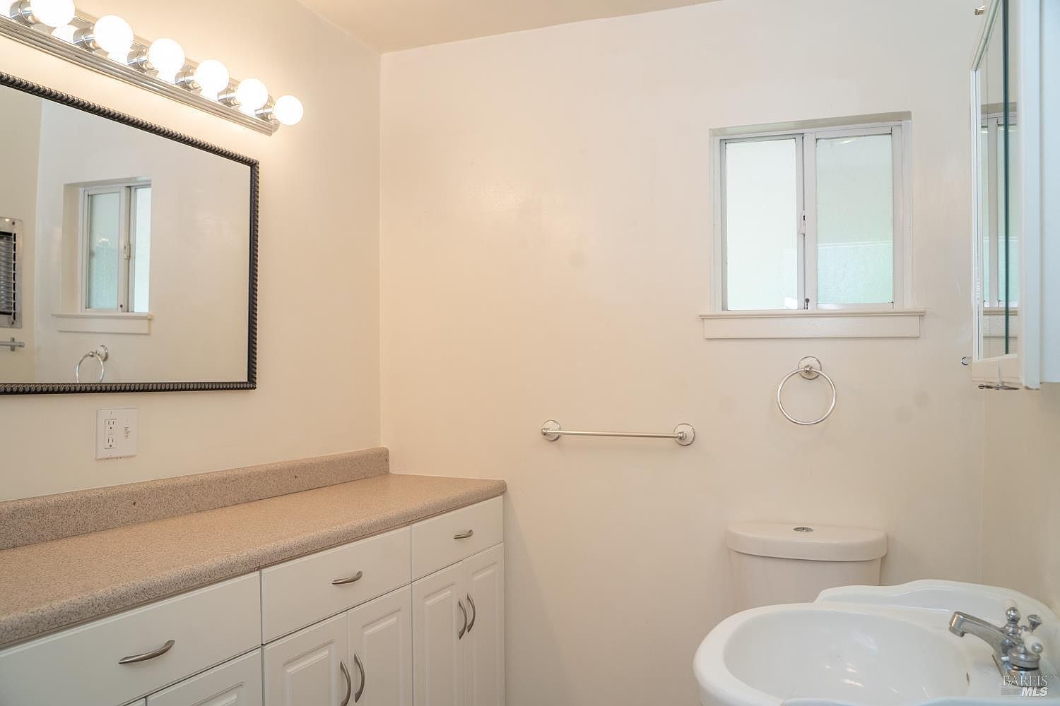 This bathroom features a neutral color palette with white cabinetry and a light-toned countertop. A large, framed mirror sits above the vanity, illuminated by a multi-bulb light fixture, while a window provides natural light to the space. The room includes a standard toilet and a wall-mounted towel ring, creating a functional and clean aesthetic.