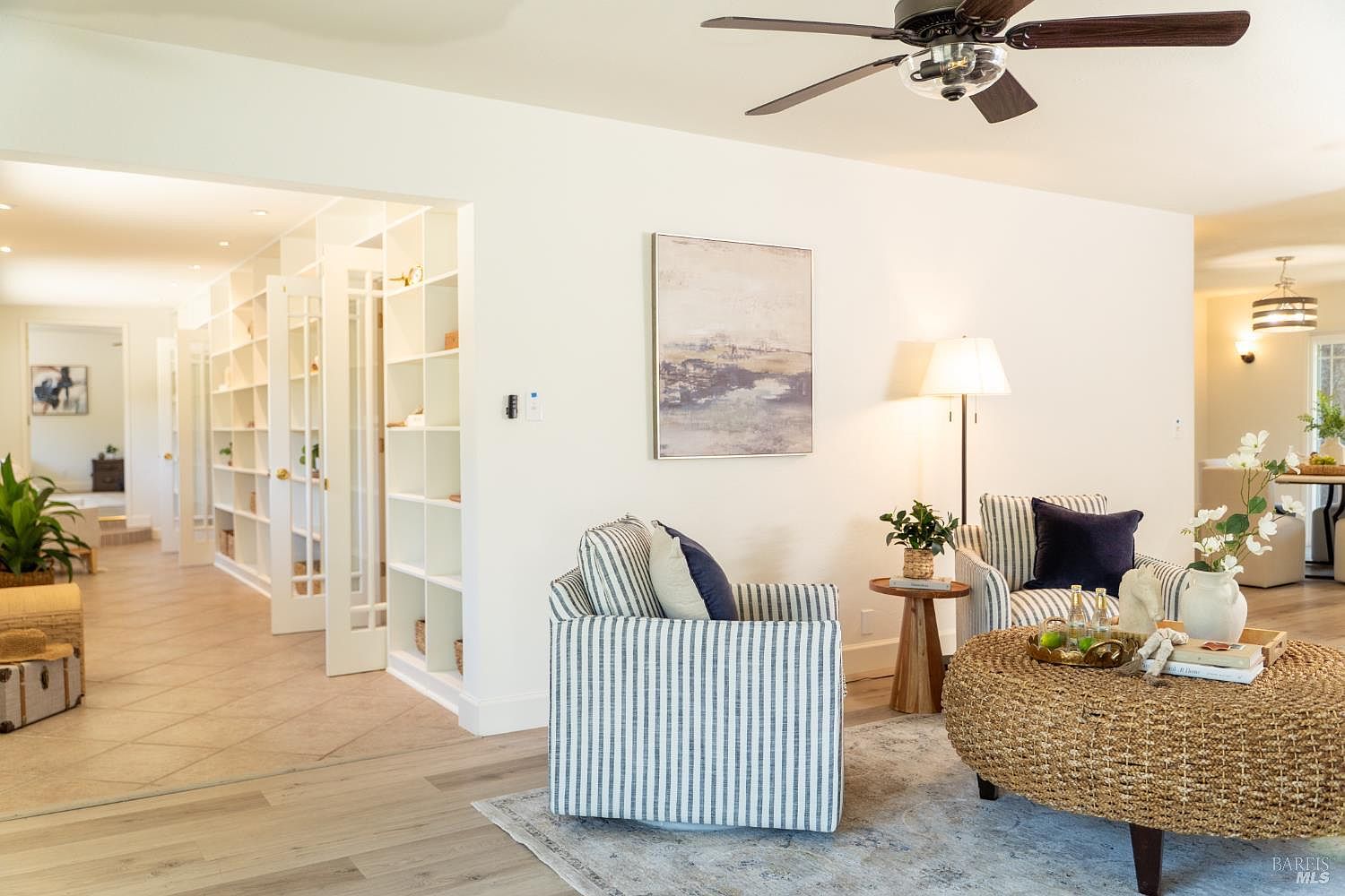 This bright and inviting living room features a pair of striped armchairs arranged around a large, round woven ottoman. To the left, a built-in shelving unit with glass-paned doors leads into a tiled hallway, while a piece of abstract art hangs on the main wall above a floor lamp. The space is finished with light-toned wood flooring and a ceiling fan, creating a cohesive and airy atmosphere.