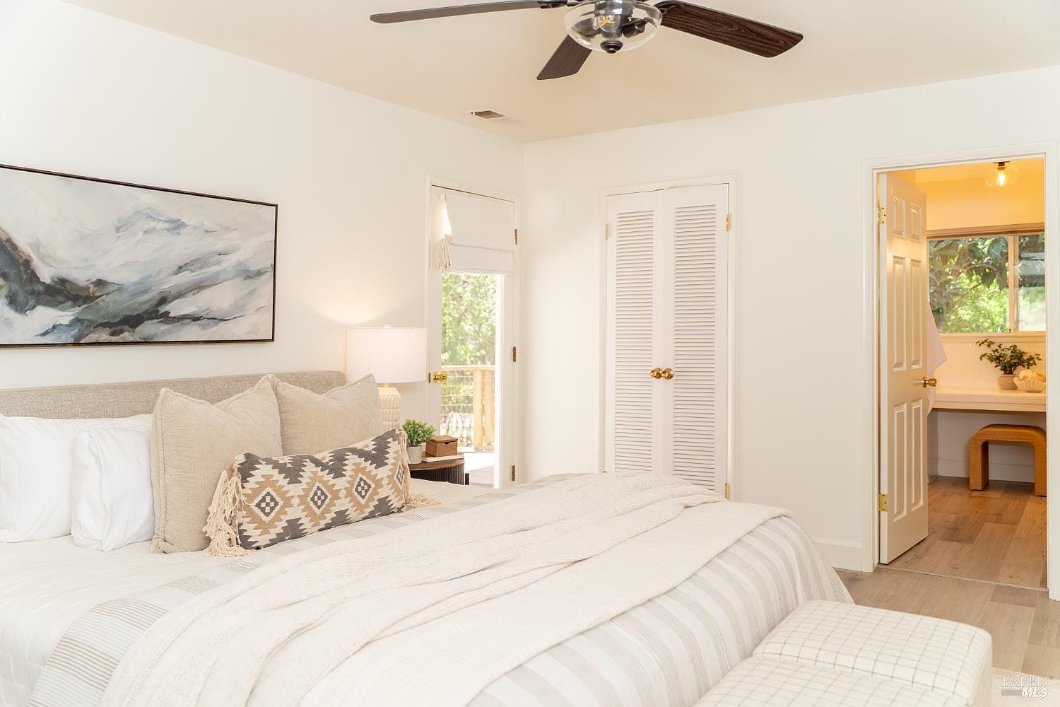 This bright and airy primary bedroom features a neutral color palette with soft textures and a comfortable bed dressed in layered linens. A large piece of abstract wall art hangs above the headboard, while a ceiling fan provides overhead lighting and circulation. The room includes a door leading to an outdoor deck and an open doorway revealing a glimpse of an adjoining bathroom with light wood flooring.