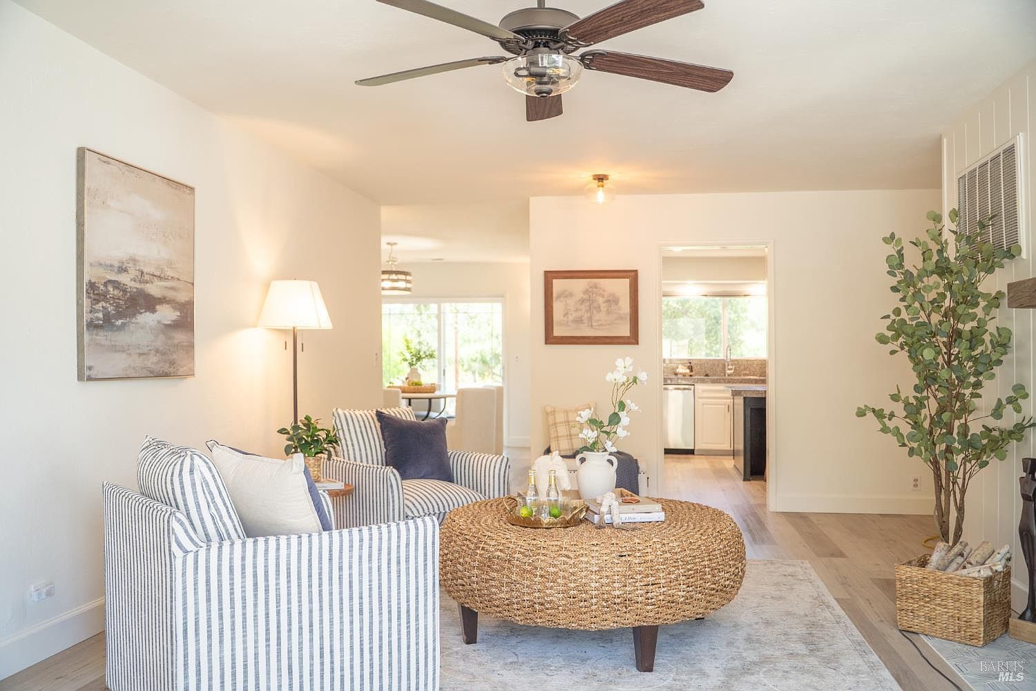 This bright and inviting living room features a pair of blue and white striped armchairs arranged around a large, round woven ottoman. The space is decorated with neutral tones, light wood-look flooring, and a large piece of abstract wall art, creating a cohesive and airy atmosphere. A view through the open doorway reveals a glimpse of the kitchen, highlighting the home's open-concept layout and natural flow.