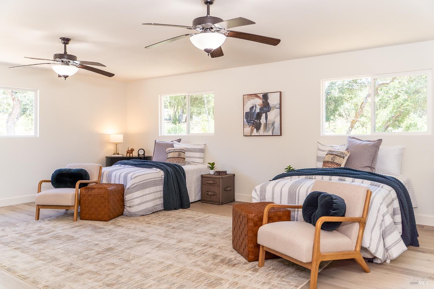 This spacious bedroom features two twin beds with striped bedding, navy blue throws, and decorative pillows, creating a cohesive and inviting aesthetic. The room is anchored by a large, neutral-toned area rug and includes two comfortable armchairs with matching ottomans, providing a cozy seating area. Large windows allow ample natural light to fill the space, while two ceiling fans provide overhead lighting and air circulation.