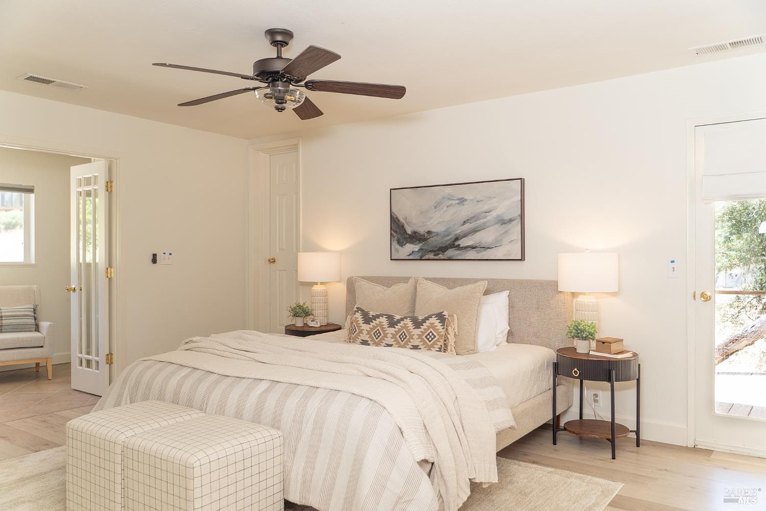 This bright and airy primary bedroom features a neutral color palette with soft textures and a modern aesthetic. The room is centered around a plush bed with layered bedding, flanked by two matching nightstands and lamps, while a pair of checkered ottomans sits at the foot of the bed. A large abstract painting hangs above the headboard, and the space is illuminated by a ceiling fan with integrated lighting, creating a calm and inviting atmosphere.