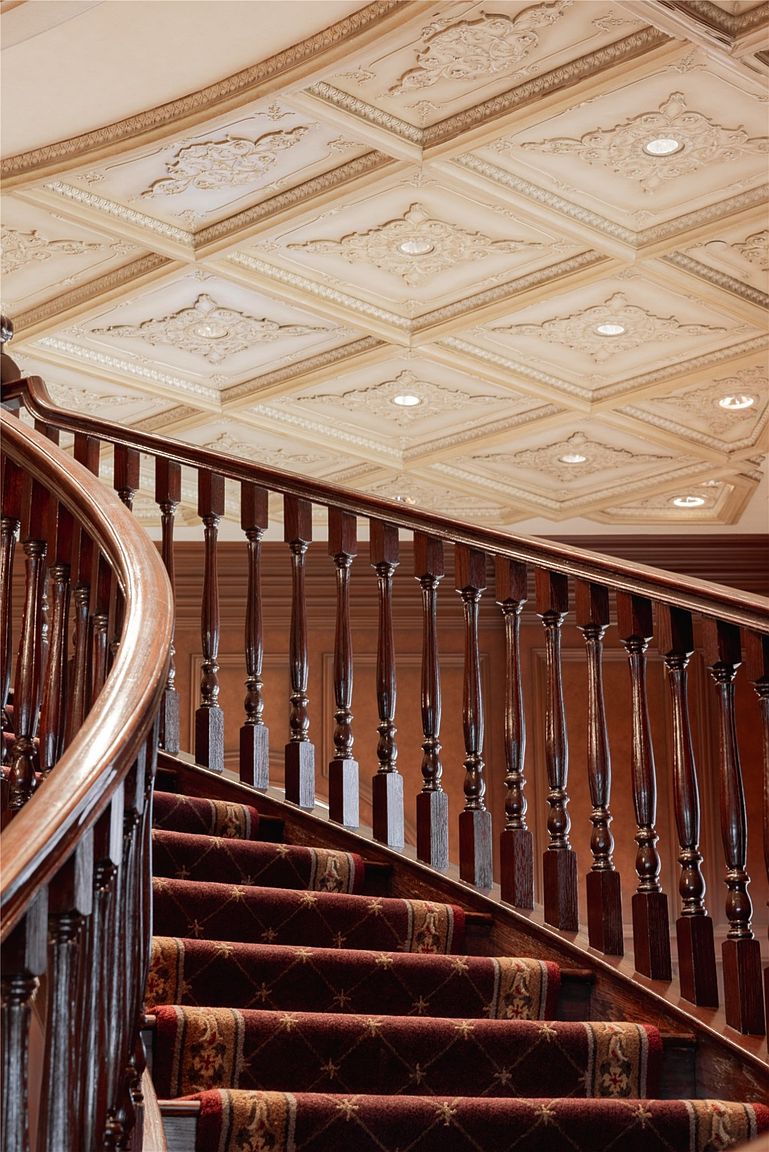 This image captures a grand, curved staircase featuring dark wood railings and balusters, complemented by a rich red patterned runner. Above, the ceiling is adorned with intricate, coffered molding and recessed lighting, creating a sophisticated and traditional atmosphere. The perspective is angled upward, emphasizing the architectural detail of the ceiling and the elegant sweep of the staircase.