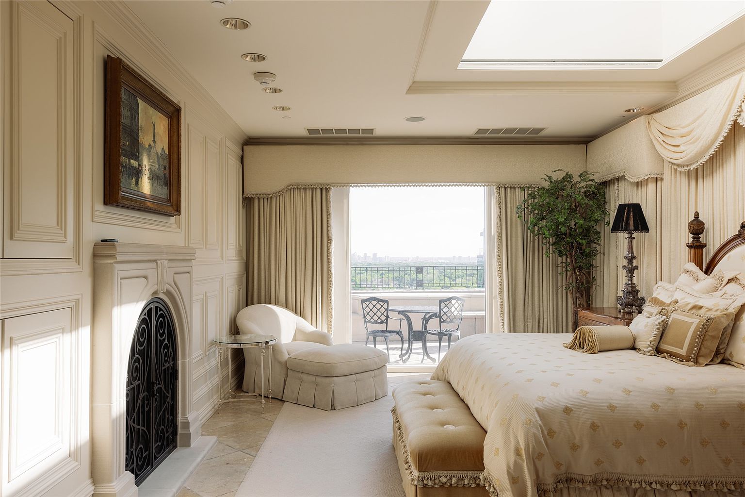 This elegant primary bedroom features a sophisticated neutral color palette, highlighted by a plush bed with ornate bedding and a matching bench at the foot. To the left, a classic fireplace with a decorative metal screen is set into a paneled wall, while a cozy chaise lounge sits near a large glass door that opens onto a private balcony. The room is bathed in natural light from both the balcony and a skylight above, creating a serene and luxurious atmosphere.