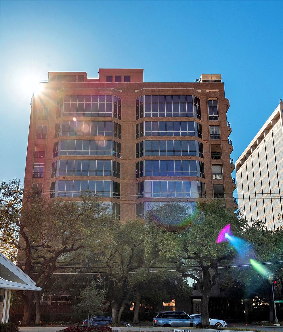 This image captures a multi-story brick residential building from a low-angle street perspective, emphasizing its height and architectural presence. The facade features symmetrical banks of large windows that reflect the bright blue sky, flanked by a vertical column of private balconies on the right side. Mature trees in the foreground partially frame the structure, while the bright sun creates a lens flare effect, adding a cinematic quality to the urban scene.