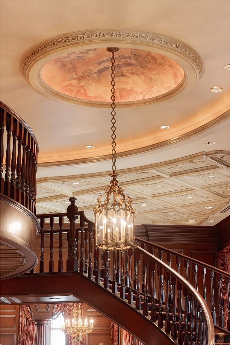This grand foyer features a sweeping wooden staircase with ornate balusters that leads to an upper level. Above, a circular ceiling medallion showcases a detailed mural, illuminated by a sophisticated hanging lantern fixture. The space is defined by rich wood paneling, intricate coffered ceiling details, and a warm, opulent atmosphere.