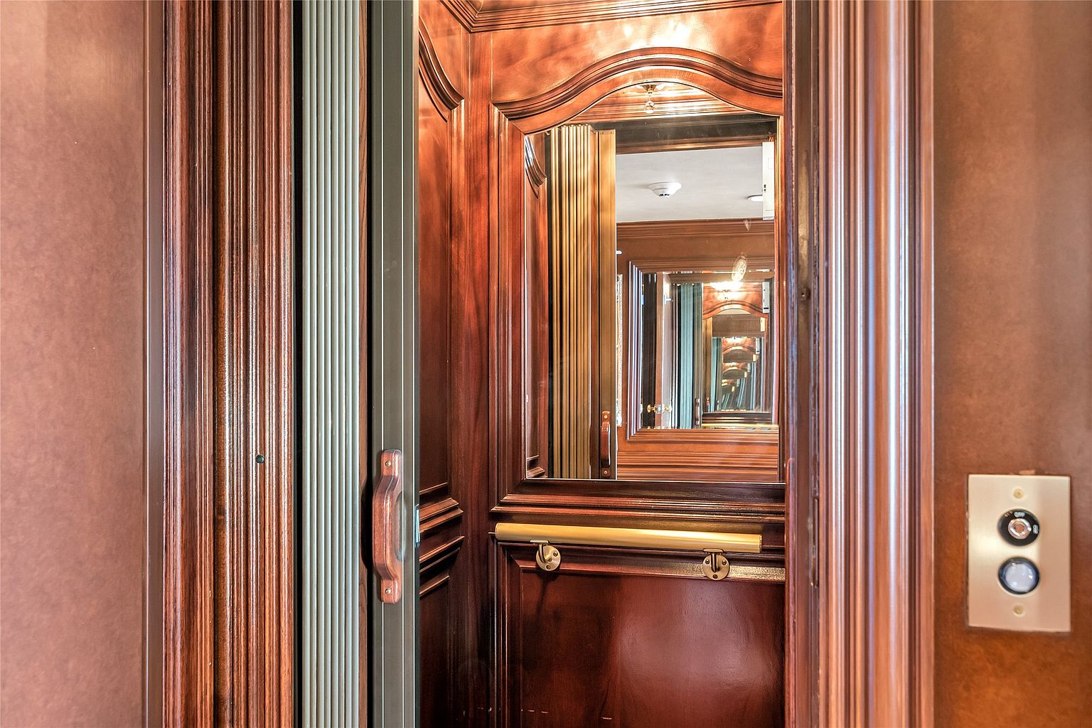 This image captures the interior of a private residential elevator, characterized by rich, dark wood paneling and classic architectural detailing. A central mirror reflects the interior space, while a brass-toned handrail and a simple control panel with two buttons are mounted on the side wall. The perspective is centered, looking directly into the elevator car, highlighting its elegant and traditional aesthetic.