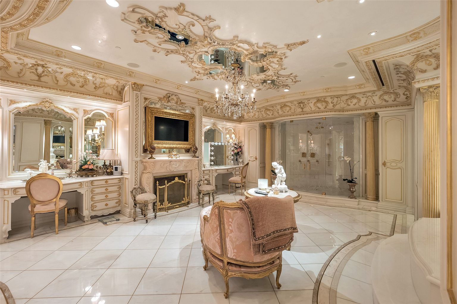 This opulent primary bathroom features an ornate, classical design with intricate gold-leaf ceiling molding, a crystal chandelier, and polished stone flooring. The space includes a vanity area with a mirror, a fireplace with a decorative mantel, and a glass-enclosed shower stall. A plush, upholstered armchair sits in the center of the room, enhancing the luxurious and sophisticated atmosphere.