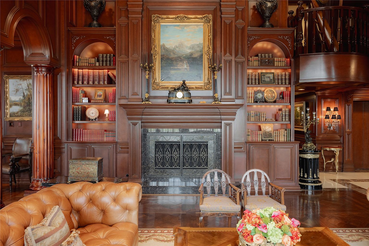 This elegant living room features rich, dark wood paneling and built-in shelving flanking a central fireplace with a decorative metal screen. A large, ornate landscape painting hangs above the mantel, complemented by classic furniture including a tufted leather sofa and two upholstered chairs. The space exudes a traditional, sophisticated atmosphere with polished wood floors and warm, ambient lighting.