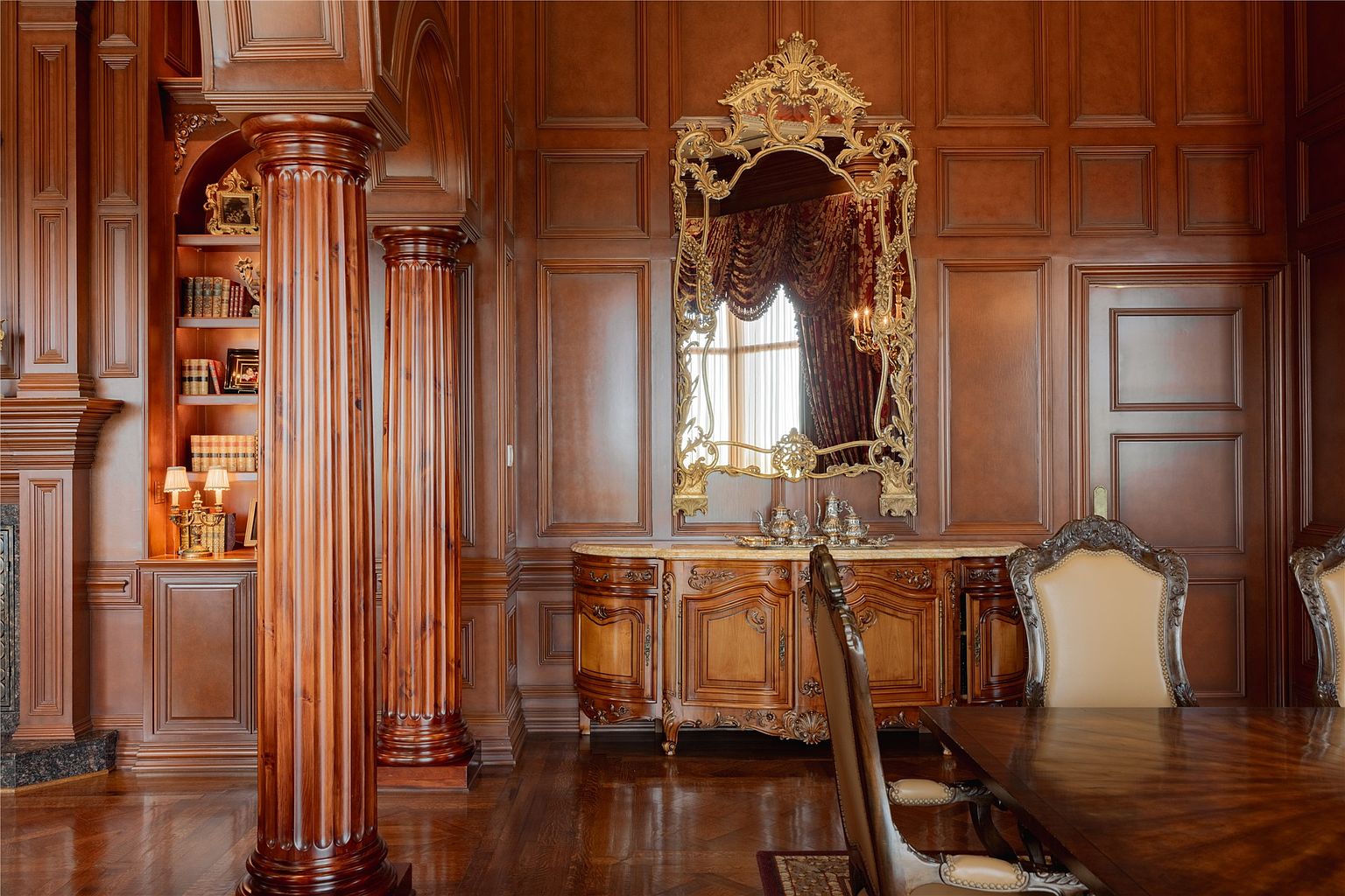 This formal dining room features rich, dark wood paneling throughout the walls, creating a sophisticated and traditional atmosphere. A large, ornate gold-framed mirror hangs above a detailed wooden sideboard, while fluted columns add architectural depth to the space. The room is furnished with a dark wood dining table and high-backed chairs, reflecting a classic and luxurious aesthetic.
