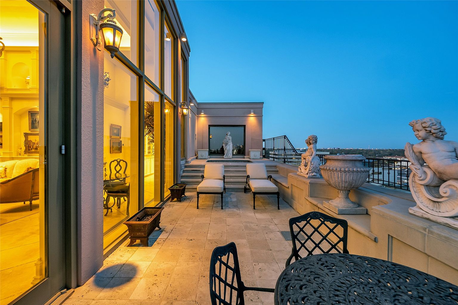 This expansive rooftop terrace offers a sophisticated outdoor living space with stone flooring and a panoramic view of the city skyline at dusk. The area is furnished with lounge chairs, a bistro set, and decorative stone statues, creating an elegant atmosphere for relaxation or entertaining. Large glass doors provide a seamless transition from the interior living space to this elevated balcony, which is illuminated by warm wall-mounted lighting.