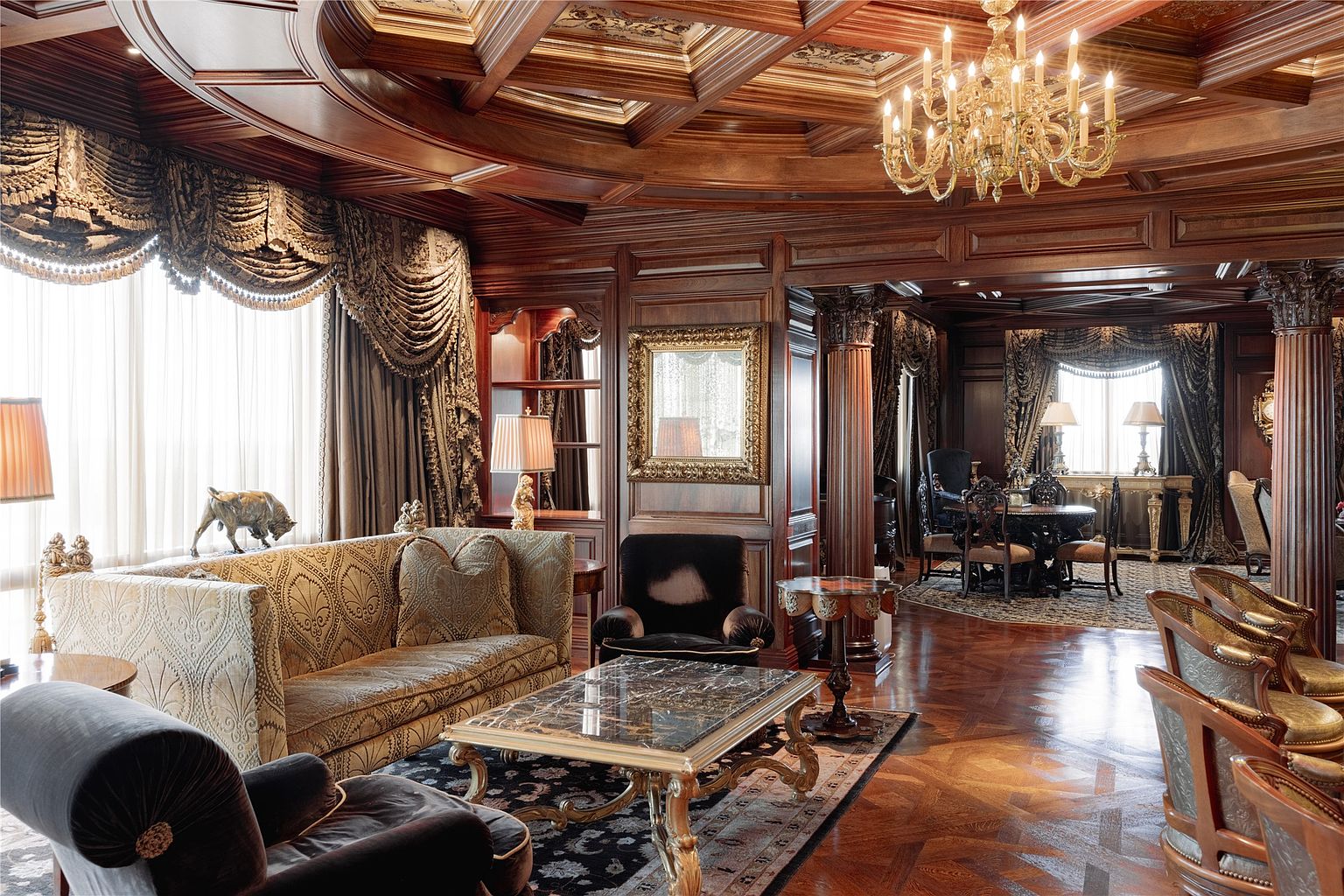 This opulent living room features rich wood-paneled walls, a coffered ceiling, and intricate parquet flooring, creating a sophisticated and traditional atmosphere. The space is furnished with a patterned sofa, plush velvet armchairs, and a stone-topped coffee table, all centered on a dark area rug. A crystal chandelier hangs prominently above, while large windows draped in heavy, ornate curtains allow natural light to fill the room, highlighting the detailed craftsmanship throughout.