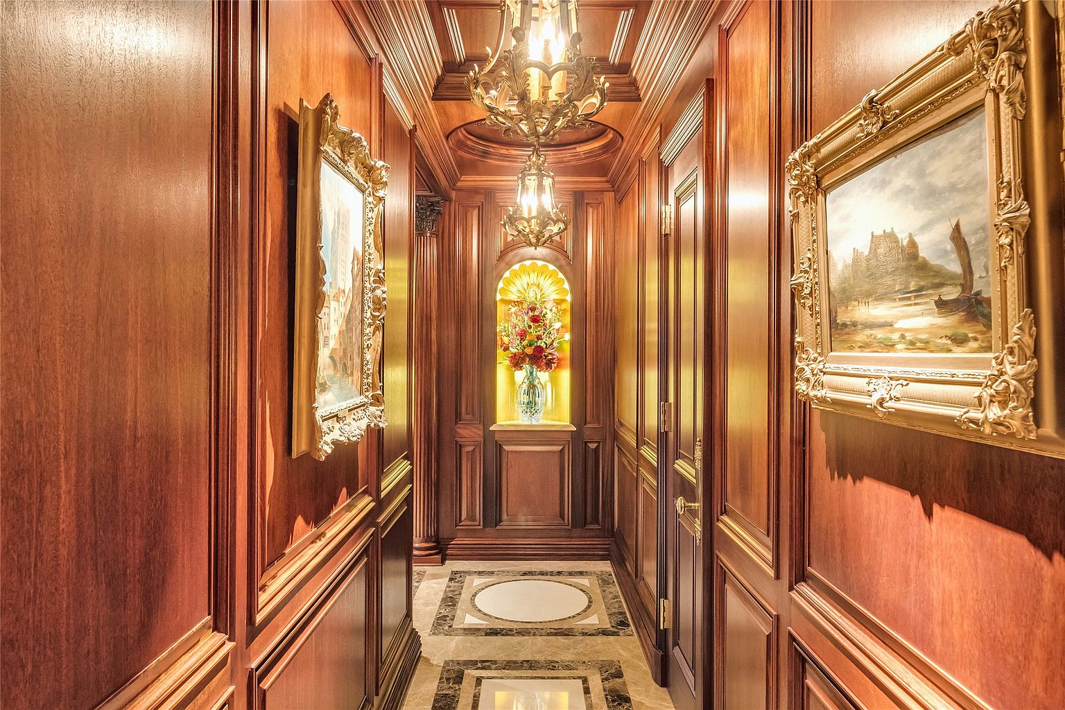 This elegant hallway features rich, dark wood paneling throughout, creating a sophisticated and traditional atmosphere. The space is illuminated by an ornate, multi-tiered chandelier and a recessed, backlit alcove displaying a floral arrangement at the end of the corridor. The floor is finished with decorative stone tiles, and the walls are adorned with framed artwork, providing a refined and gallery-like aesthetic.