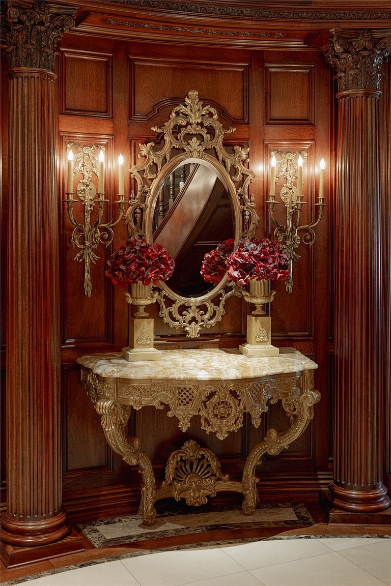 This elegant vignette features a highly ornate, gold-toned console table topped with a polished stone surface, positioned against rich, dark wood paneling. Above the table hangs an intricate, matching gold-framed oval mirror flanked by two classic wall-mounted candelabra sconces. Two decorative urns filled with deep red floral arrangements complete this sophisticated and formal display.
