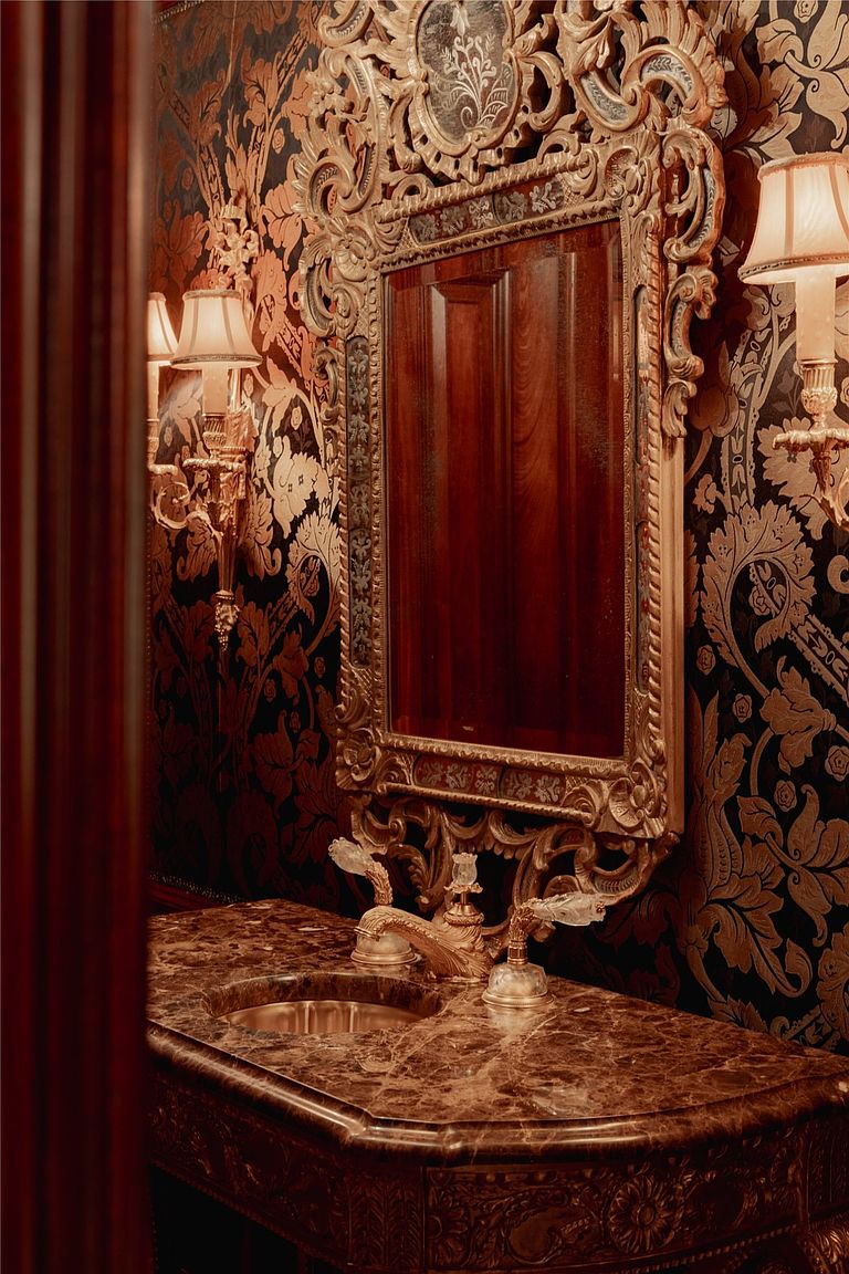 This opulent powder room features a dark, ornate damask wallpaper and a heavy, gold-gilded mirror frame that serves as a dramatic focal point. A rich, dark stone vanity with a polished finish supports a decorative faucet, while matching wall sconces provide warm, ambient lighting. The overall aesthetic is one of classic, high-end luxury with a moody and sophisticated atmosphere.