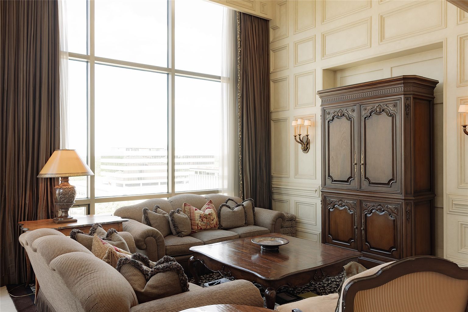 This elegant living room features a sophisticated traditional design with paneled walls and tall windows that allow for abundant natural light. The space is furnished with plush, neutral-toned sofas arranged around a substantial dark wood coffee table, creating a comfortable and inviting seating area. A large, ornate wooden armoire serves as a focal point against the wall, complemented by warm wall sconce lighting and rich, floor-length drapery.