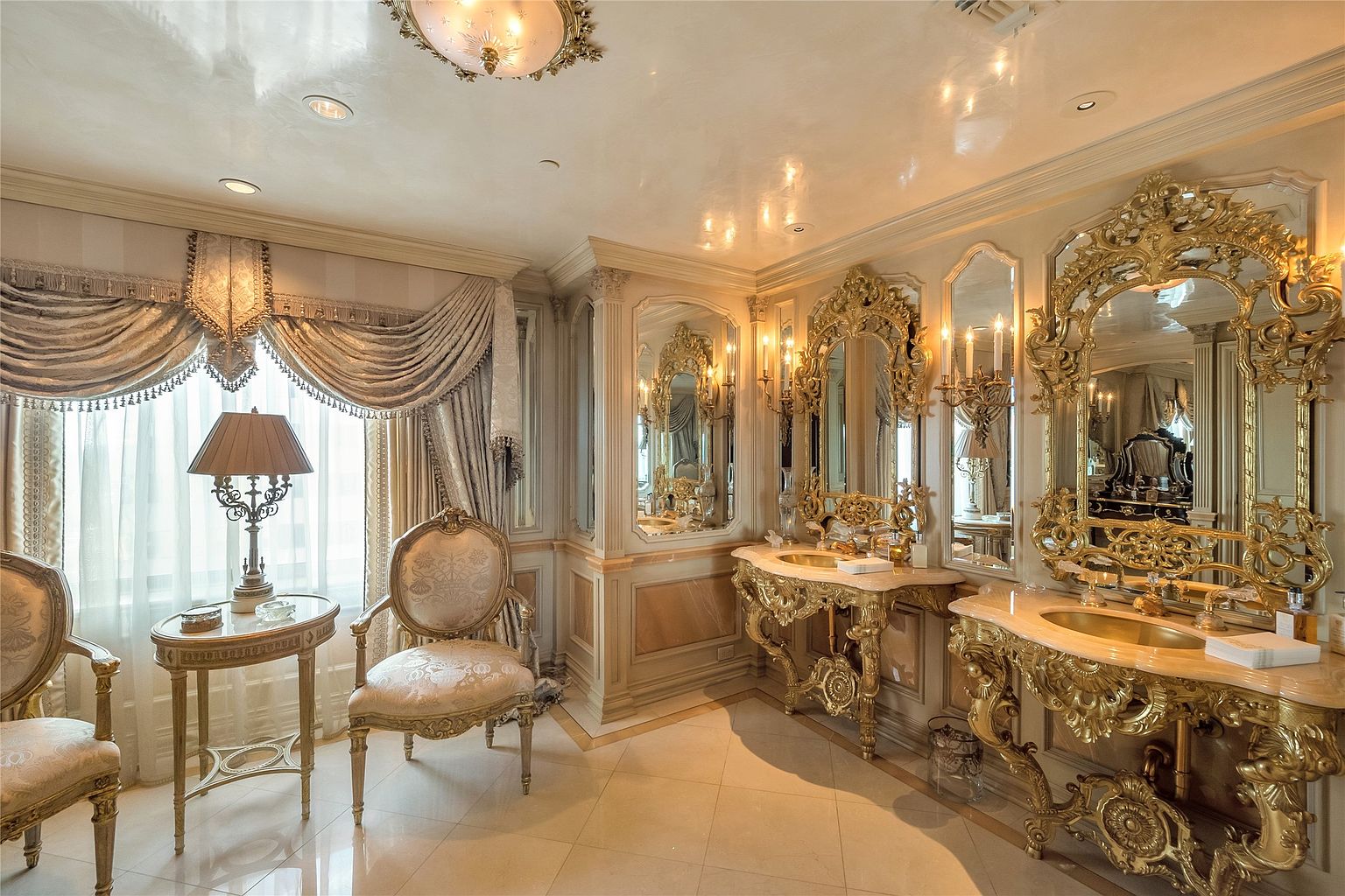 This opulent primary bathroom features an ornate, gold-leaf vanity with dual sinks and intricate, carved detailing beneath large, gilded mirrors. The room is elegantly appointed with a seating area consisting of two upholstered chairs and a small side table, all framed by heavy, draped window treatments. The space is finished with polished stone flooring and a decorative ceiling fixture, creating a highly luxurious and classical aesthetic.