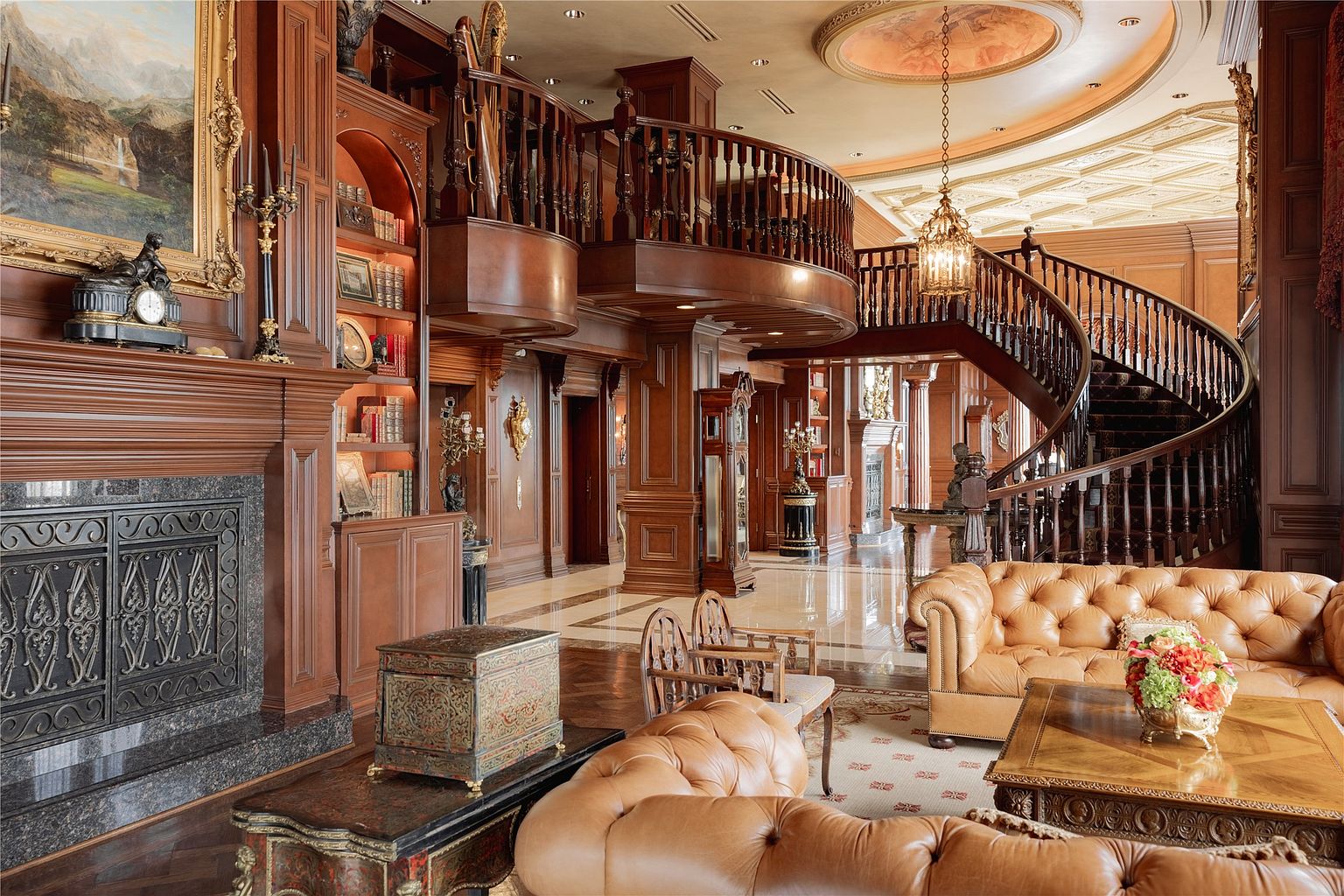 This grand, opulent living room features rich wood paneling, a sweeping curved staircase, and a mezzanine balcony overlooking the space. The room is furnished with tufted leather sofas and ornate decor, including a large painting above a stone-surround fireplace and a decorative ceiling medallion with a chandelier. The perspective captures the expansive, multi-level layout, emphasizing the luxurious, traditional architectural details and the polished stone flooring.
