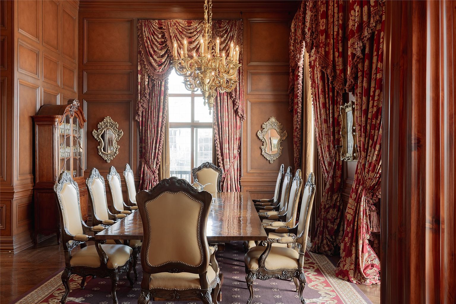 This formal dining room exudes traditional elegance with its rich wood-paneled walls and ornate, heavy drapery framing a large window. A grand crystal chandelier serves as the centerpiece above a long, polished dining table surrounded by high-backed, upholstered chairs. The space is further accented by decorative wall mirrors and a classic china cabinet, creating a sophisticated and opulent atmosphere.