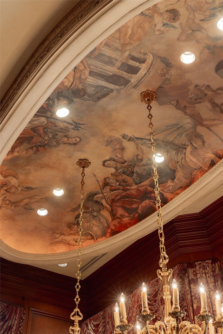 This image captures a dramatic, oval-shaped recessed ceiling featuring an elaborate, classical-style mural depicting mythological figures and architectural motifs. The ceiling is illuminated by multiple recessed lights and accented by a pair of ornate, gold-toned chandeliers hanging from the center. Below the ceiling, the room features rich, dark wood paneling and heavy, patterned drapery, creating a sophisticated and opulent atmosphere.