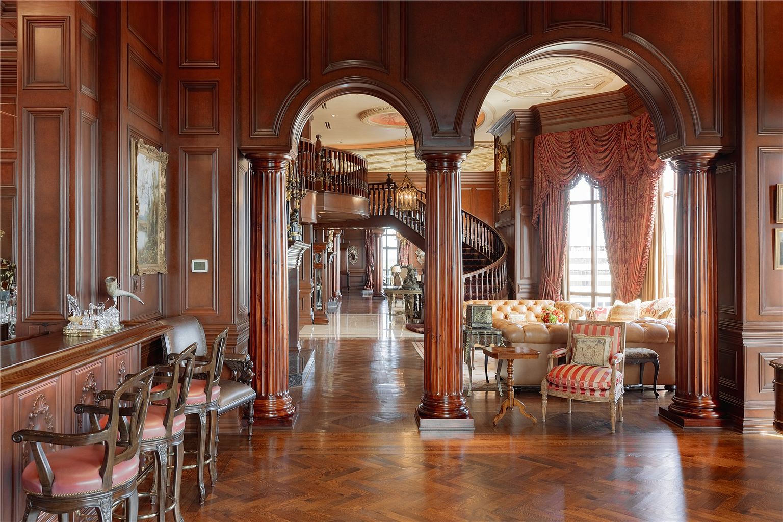 This grand interior space features rich, dark wood paneling throughout, creating a sophisticated and traditional atmosphere. The perspective looks through a series of arched openings supported by fluted columns, leading toward a sweeping curved staircase in the background. To the right, a formal seating area with a tufted leather sofa and patterned armchair is visible, while a bar area with stools occupies the foreground.