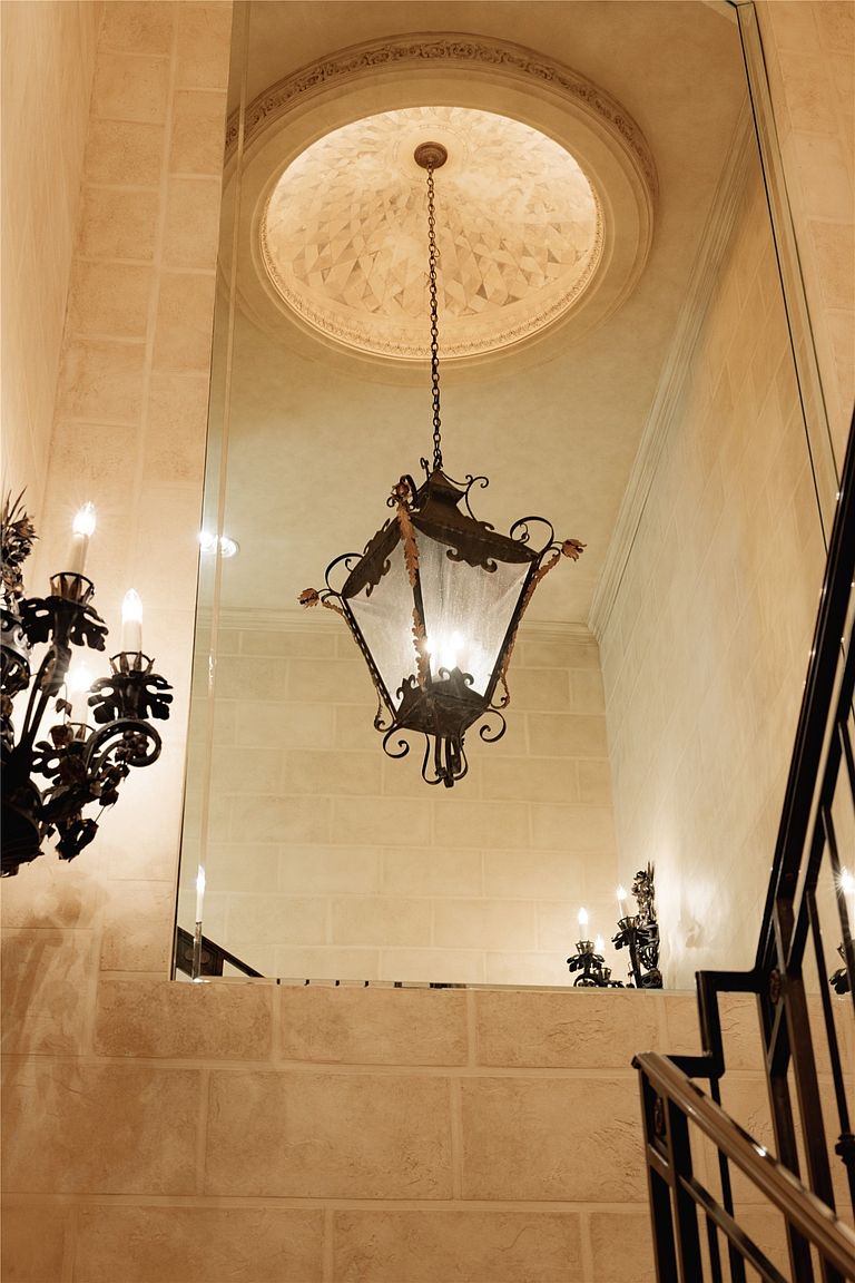 This upward-looking perspective captures a grand staircase area featuring a dramatic, ornate metal lantern pendant light suspended from a recessed, circular domed ceiling. A large, seamless mirror reflects the space, creating an illusion of depth and highlighting the textured, light-colored stone wall finish. The scene is accented by a decorative wall sconce and a dark metal railing, contributing to an elegant, Old World architectural aesthetic.