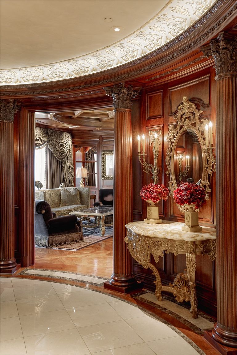 This elegant entryway features rich wood-paneled walls, ornate columns, and a decorative gold console table topped with floral arrangements and a mirror. The perspective looks through a grand arched opening into a formal living area, showcasing a sophisticated, traditional design aesthetic. The space is defined by its intricate ceiling molding and polished light-colored flooring, creating a luxurious and welcoming atmosphere.
