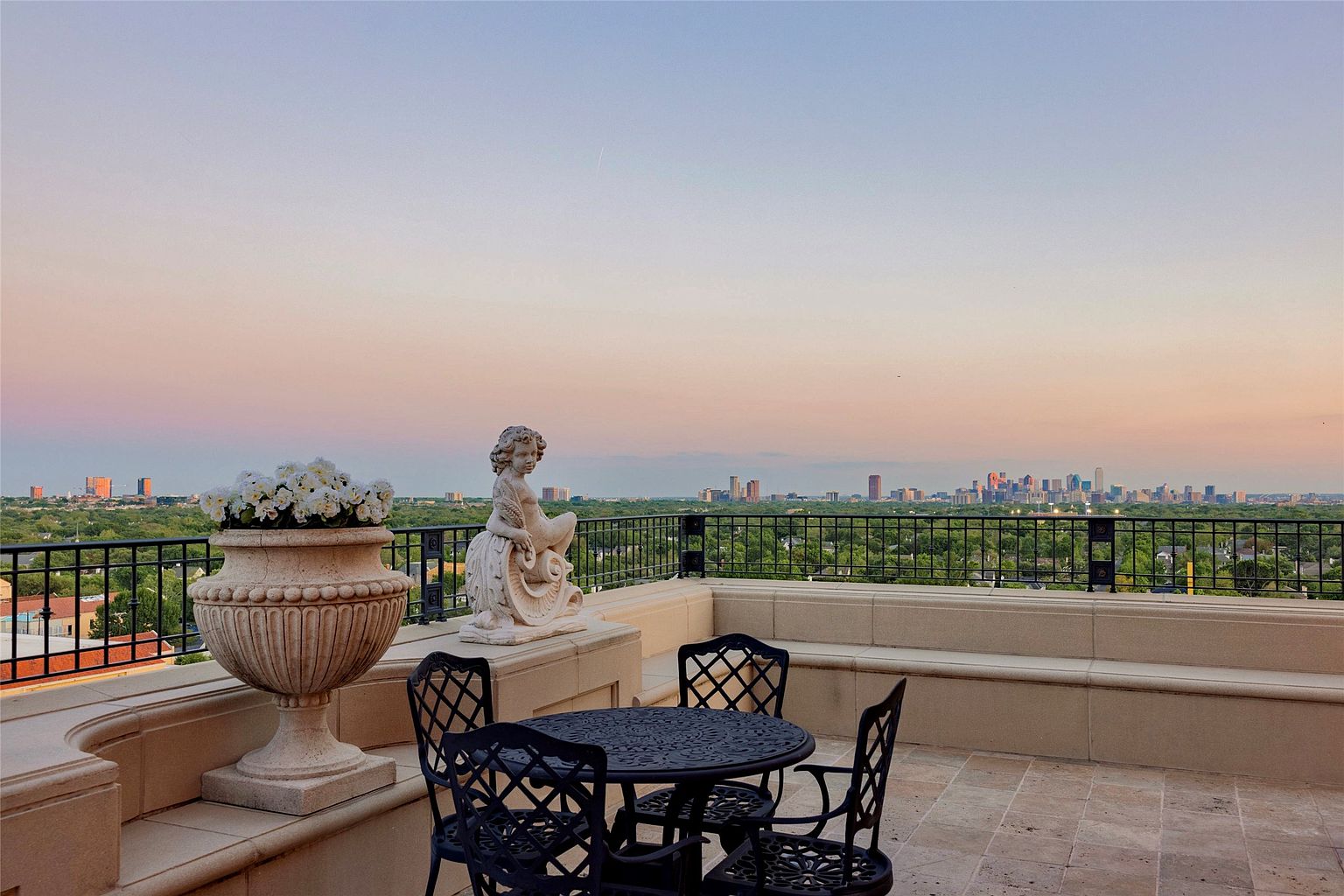 This elevated balcony offers a sophisticated outdoor living space with a panoramic view of the city skyline at sunset. The area is furnished with a dark metal bistro set and features a decorative stone planter filled with white flowers alongside a classical-style cherub statue. The scene is framed by a black wrought-iron railing, creating an elegant and serene atmosphere perfect for evening relaxation.