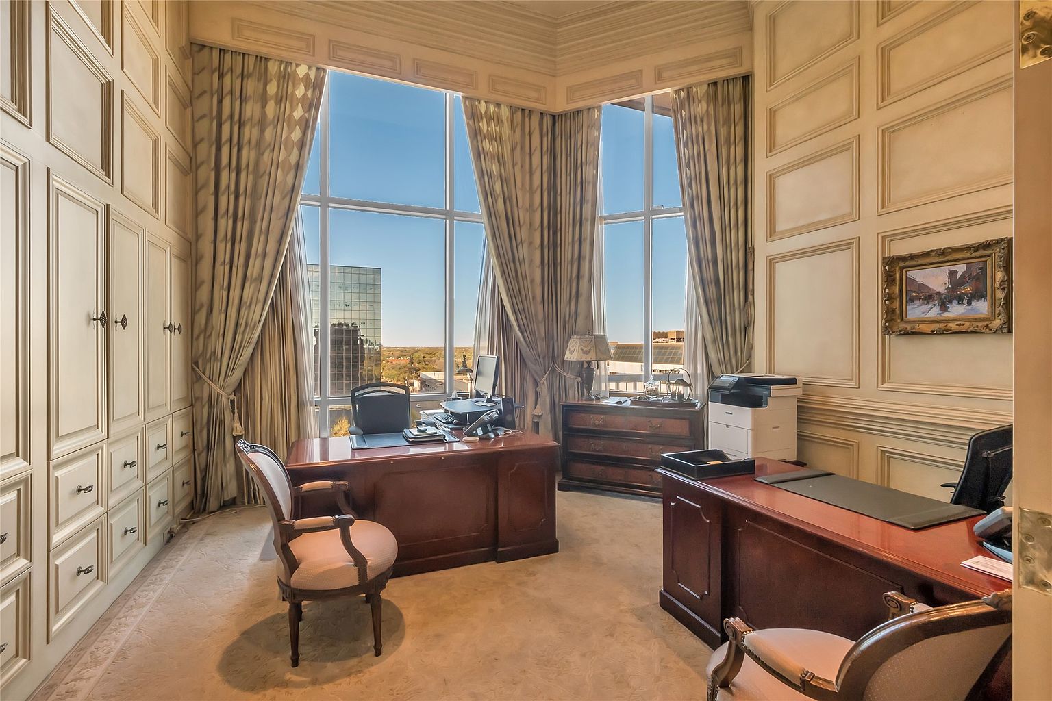 This sophisticated home office features extensive custom wall paneling and built-in cabinetry, creating a formal and professional atmosphere. The room is anchored by two large, dark wood desks and is bathed in natural light from tall windows that offer an expansive city view. Elegant drapery and classic furniture pieces complete the refined aesthetic of this executive workspace.