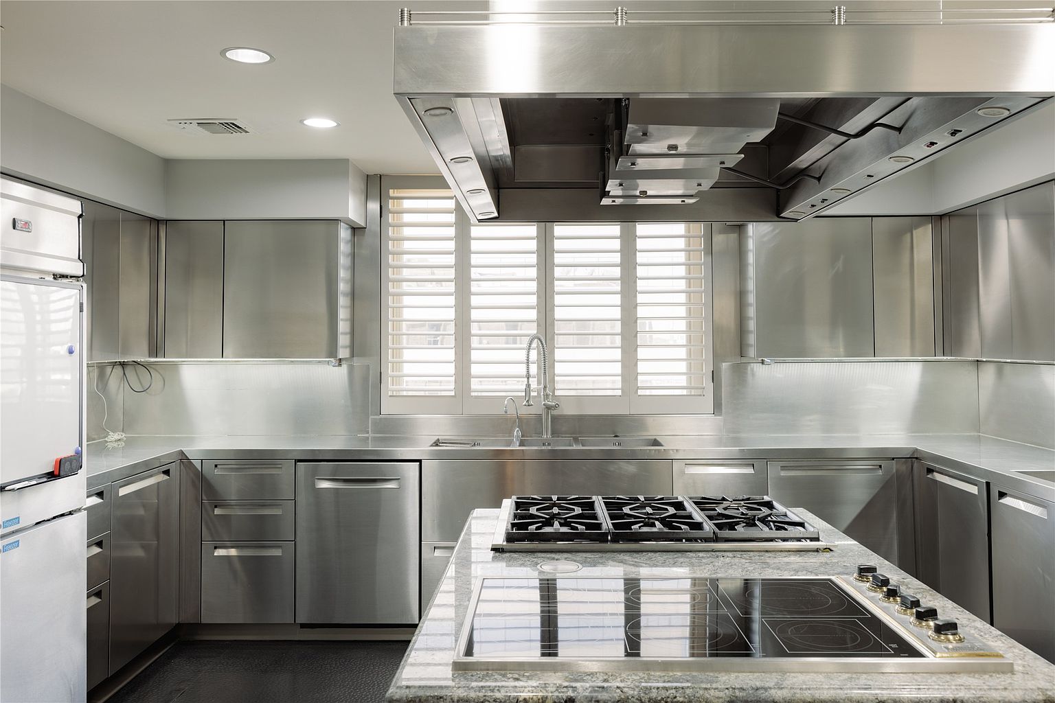 This professional-grade kitchen features extensive stainless steel cabinetry and matching metal countertops, creating a sleek, industrial aesthetic. A large central island serves as the focal point, equipped with both a gas cooktop and an electric induction range, positioned directly beneath a substantial stainless steel range hood. The space is illuminated by recessed lighting and natural light filtering through plantation shutters over the sink, offering a highly functional and modern culinary environment.