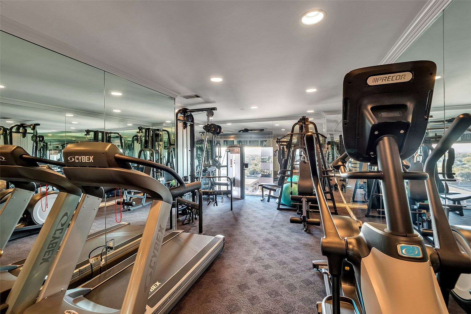 This well-equipped home gym features a variety of professional-grade fitness equipment, including Cybex treadmills and a Precor elliptical machine. The room is lined with large mirrors that enhance the sense of space, while a window at the far end provides natural light and a view of the surrounding area. The space is finished with dark carpeting and recessed ceiling lighting, creating a functional and focused workout environment.