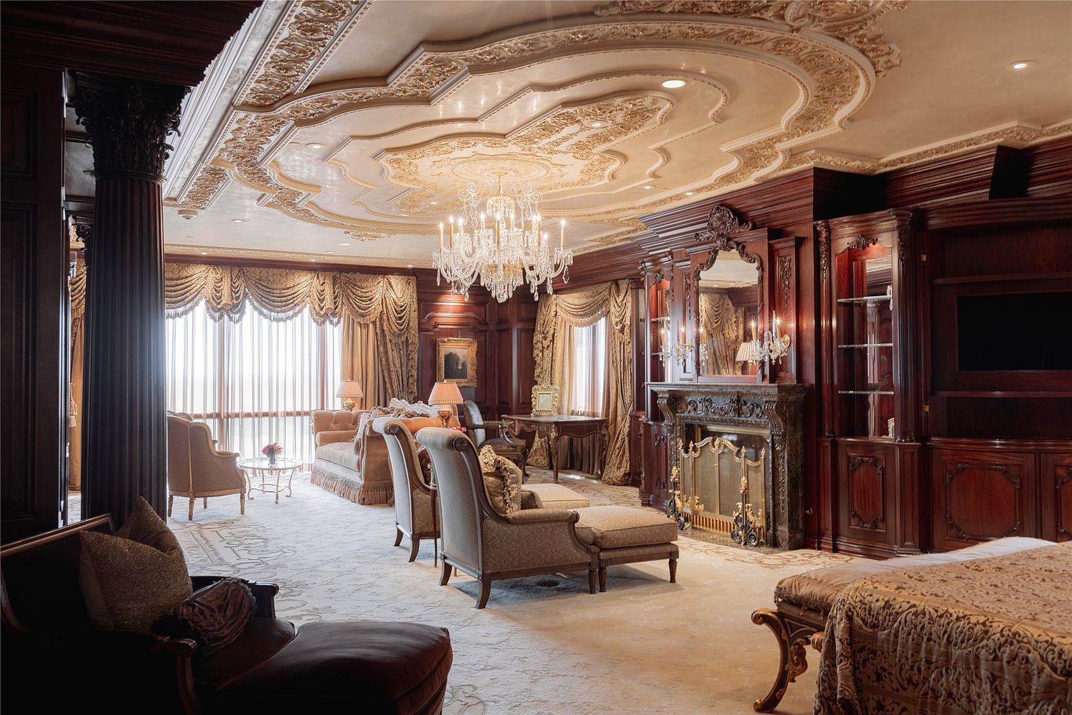 This opulent primary bedroom features an ornate, coffered ceiling with intricate gold-leaf detailing and a grand crystal chandelier. The room is anchored by a dark wood-paneled wall housing a fireplace with a decorative mantel and a built-in media center. Large windows draped in heavy, layered fabric treatments allow natural light to fill the space, which is furnished with plush seating and a patterned area rug.