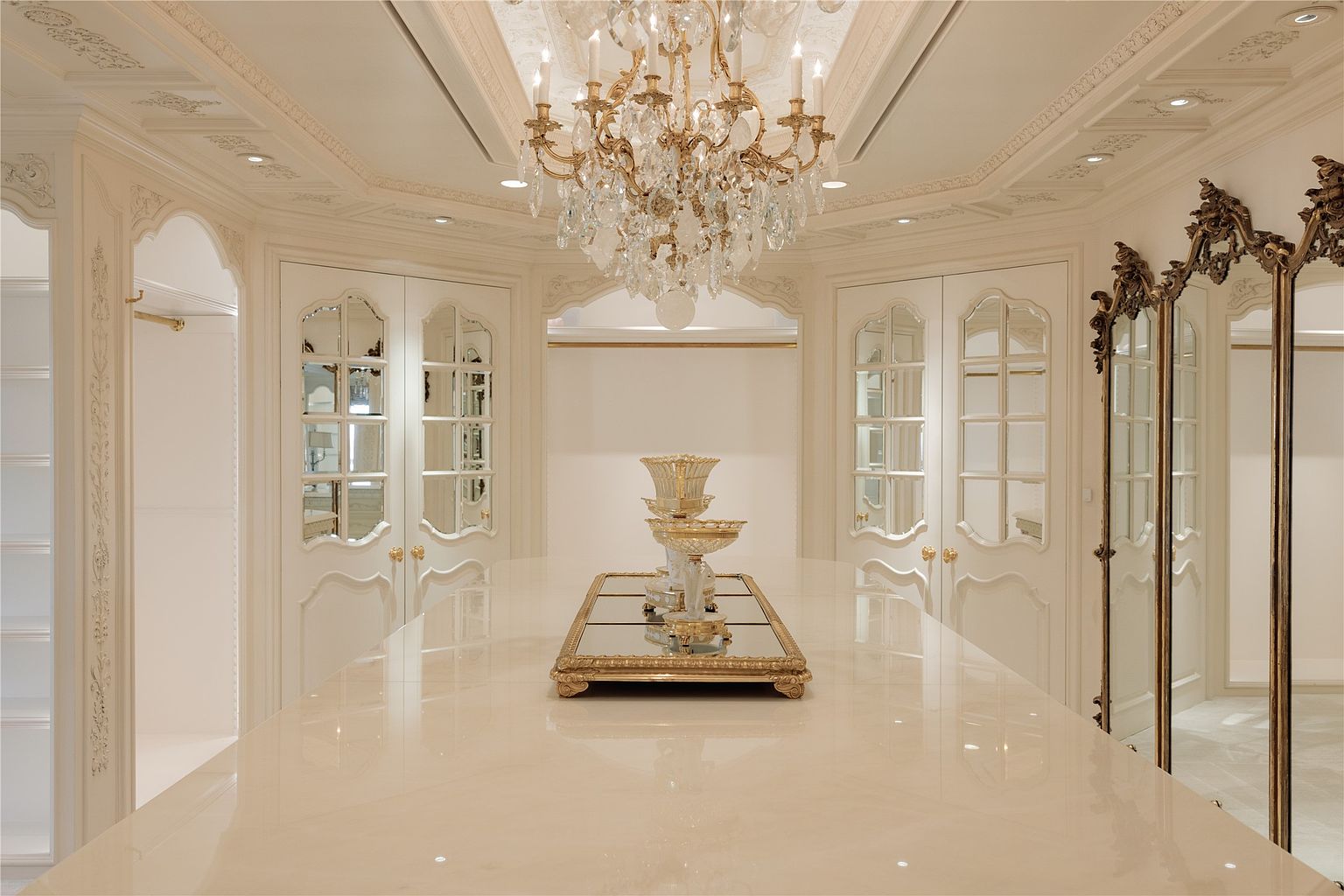 This luxurious walk-in closet features a central island with a polished stone surface and an ornate decorative centerpiece. The room is illuminated by a grand crystal chandelier hanging from a detailed ceiling, while mirrored cabinet doors and a large, gold-framed floor mirror enhance the sense of space and elegance. The perspective is centered, showcasing the symmetrical layout and sophisticated, classic design of the dressing area.