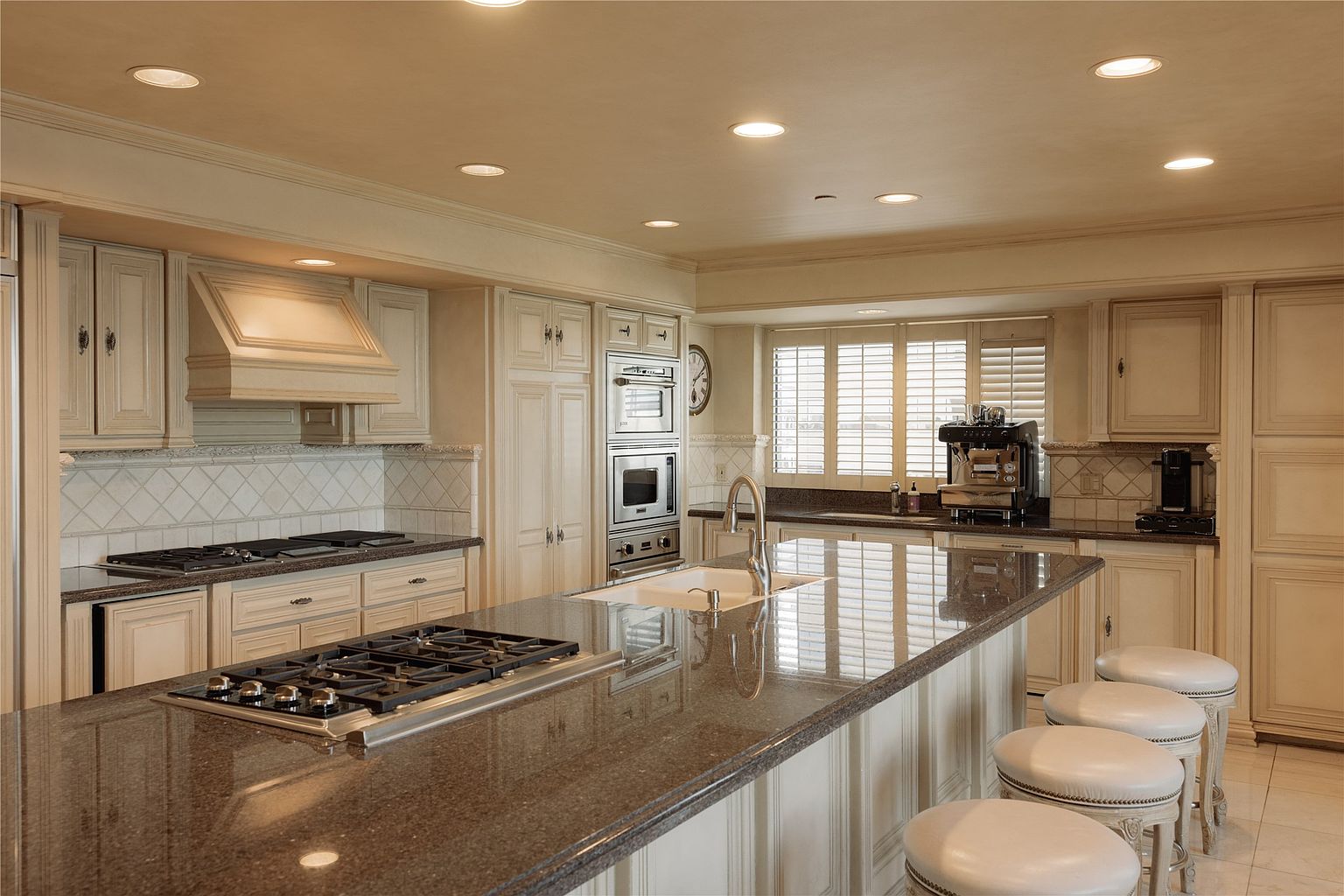 This spacious, traditional-style kitchen features light-toned cabinetry, dark stone countertops, and a large central island equipped with a gas cooktop and a secondary sink. The room is illuminated by recessed lighting and includes high-end stainless steel appliances, such as a built-in oven and microwave, alongside a row of four upholstered bar stools. The perspective captures the functional layout of the workspace, highlighting the contrast between the light cabinetry and the dark, polished stone surfaces.