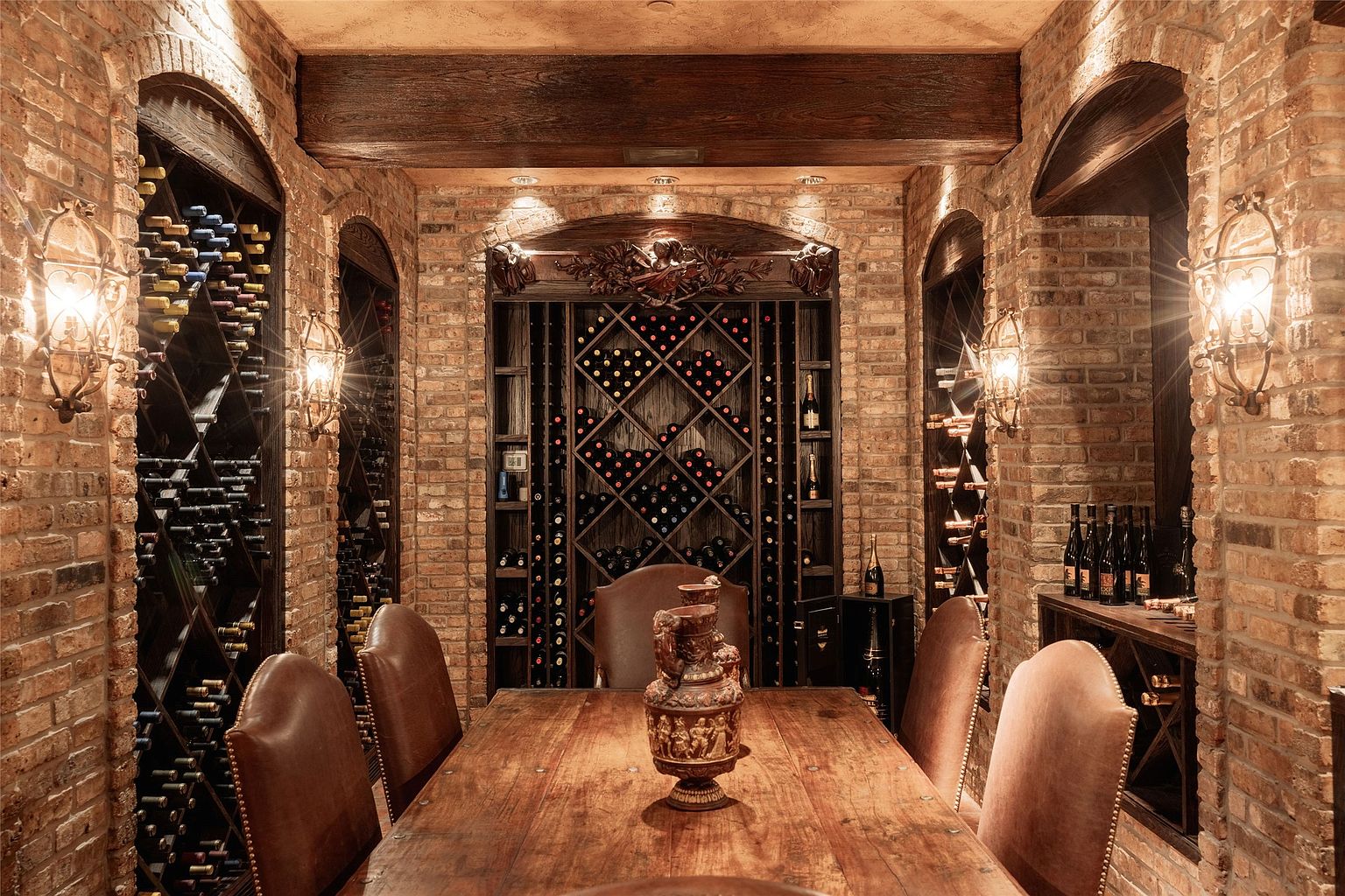 This sophisticated wine cellar features rustic brick walls and a heavy timber ceiling beam, creating an intimate, old-world atmosphere. A long wooden table sits in the center, surrounded by leather chairs and topped with an ornate decorative urn, while extensive custom wine racking lines the walls. The space is illuminated by warm, wall-mounted lanterns that highlight the textured masonry and the collection of wine bottles.