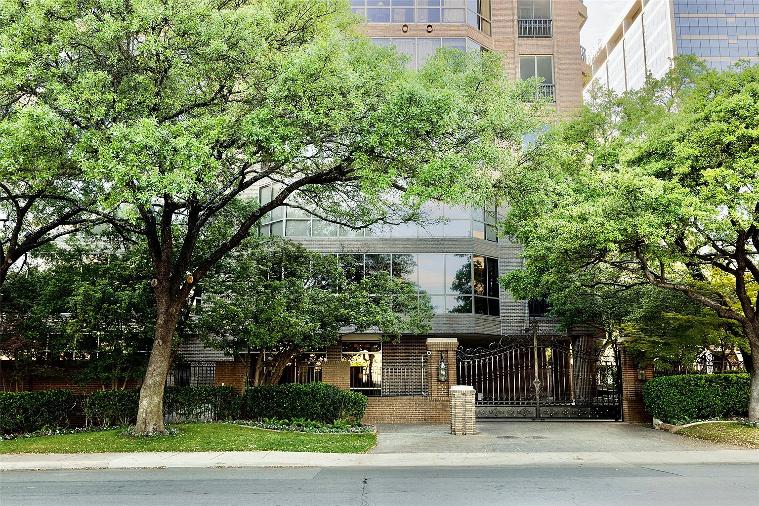 This image captures the front facade of a multi-story brick residential building, framed by mature, lush green trees. The structure features large, reflective windows and a gated entrance with a decorative metal fence, suggesting a secure and private urban residence. The perspective is a straight-on, eye-level shot that emphasizes the building's architectural presence and its integration with the surrounding greenery.