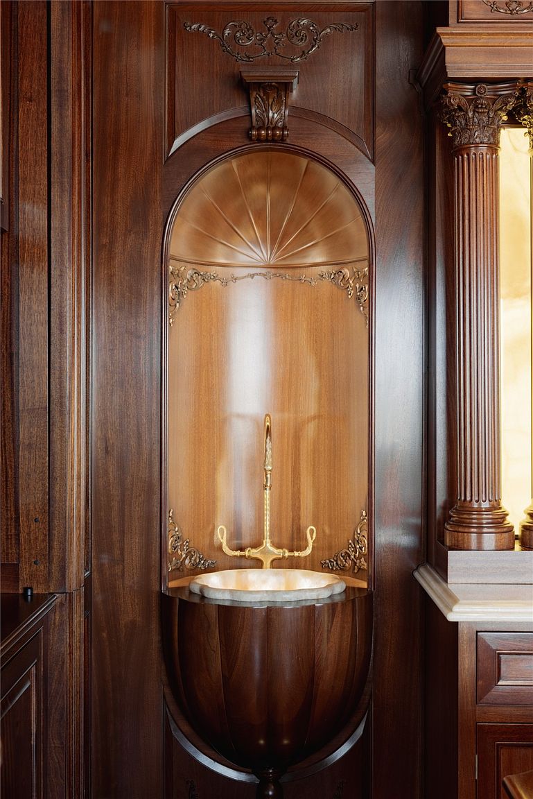 This image showcases a highly ornate, built-in wall fountain or sink feature set within a dark wood-paneled alcove. The design features a scalloped, shell-like basin, a central gold-toned faucet, and intricate decorative carvings, all framed by a curved, sunburst-patterned wood arch. The composition is centered and symmetrical, highlighting the craftsmanship and luxurious, traditional aesthetic of the space.