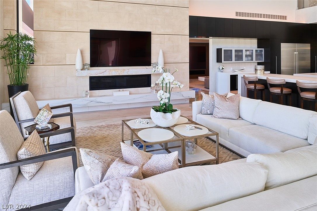 This is an interior shot of a luxurious living room featuring a modern fireplace with a large flat-screen TV above it. The room is furnished with comfortable white sofas, stylish armchairs, and a unique coffee table, creating an inviting and sophisticated atmosphere. The open floor plan seamlessly connects to the kitchen area, enhancing the sense of spaciousness and modern living.