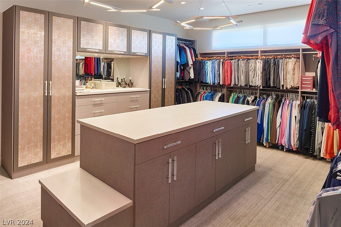 This is a spacious walk-in closet featuring custom cabinetry with a light brown wood grain finish and light countertops. The closet includes ample hanging space for clothes, drawers, and a central island with additional storage. Modern lighting fixtures illuminate the space, creating a luxurious and organized feel.