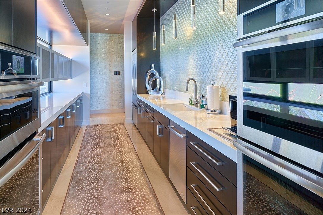This is a modern kitchen with sleek, dark cabinetry and stainless steel appliances, including double ovens. A long countertop runs along one side with a sink and decorative elements, while a patterned runner adds texture to the floor. The overall impression is one of contemporary elegance and functionality.