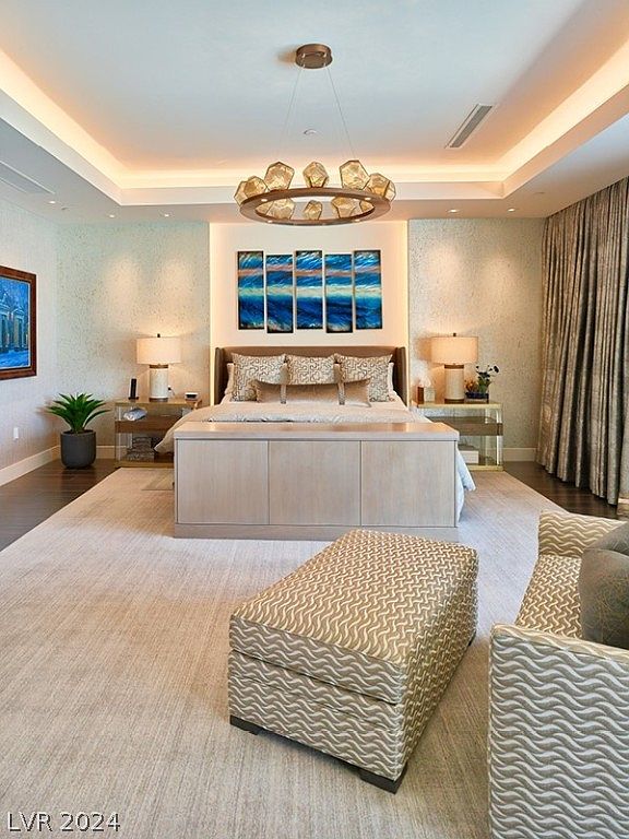 This is a primary bedroom featuring a large bed with decorative pillows and a unique headboard. The room is illuminated by a modern chandelier and recessed lighting, creating a warm and inviting ambiance. The neutral color palette and textured walls contribute to a sophisticated and relaxing atmosphere.