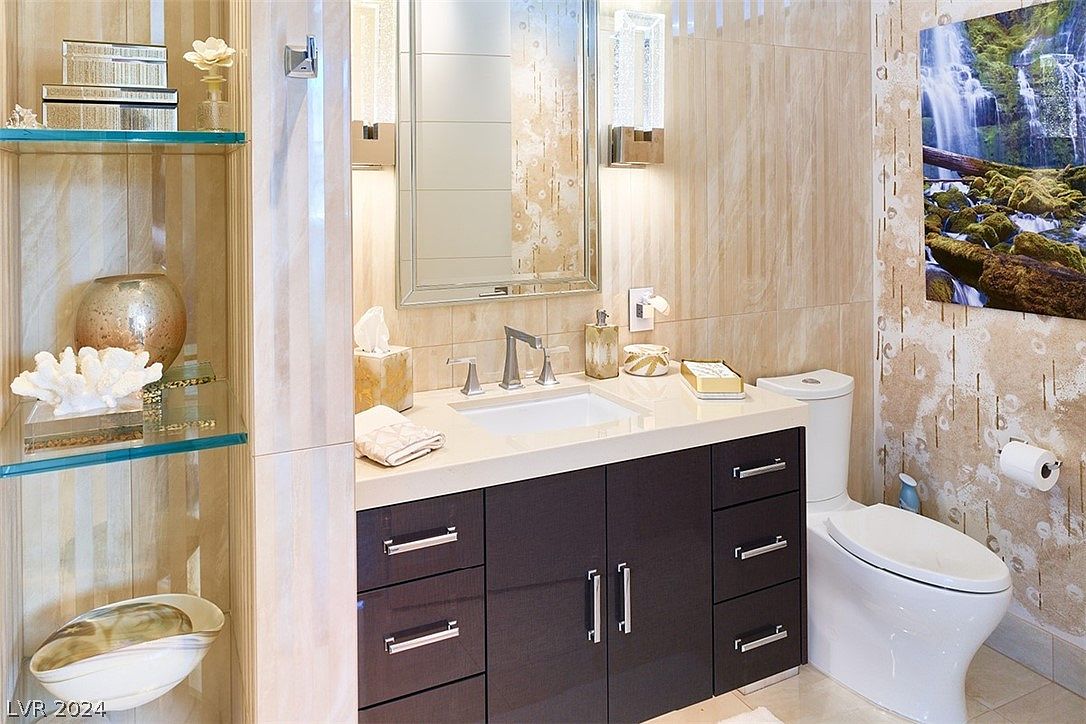 This is a well-appointed bathroom featuring a dark wood vanity with a white countertop and a rectangular sink. A decorative mirror hangs above the vanity, flanked by sconce lighting. A toilet is positioned to the right of the vanity, and a waterfall art piece hangs on the wall. The overall style is modern and luxurious.
