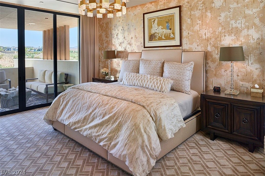 This is a luxurious primary bedroom featuring a large bed with plush bedding and a padded headboard. The room is decorated with elegant wallpaper, a stylish chandelier, and a nightstand with a lamp. A sliding glass door leads to a balcony, offering a view of the landscape.